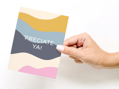 A hand with manicured nails holds the Onderkast Studio ’Preciate Ya Greeting Card, featuring wavy gold, yellow, blue, gray, cream, and pink stripes and “’PRECIATE YA!” in white. Printed in the USA.