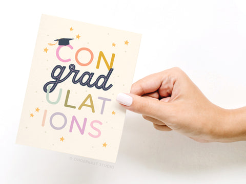 A hand with light pink nail polish holds the Onderkast Studio ConGradulations Greeting Card, featuring a graduation cap on the “C” and colorful stars—perfect for celebrating academic achievements.