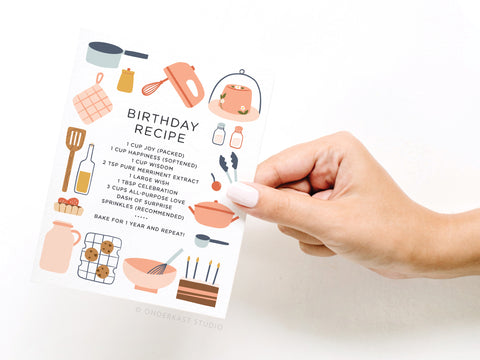A hand holds the Onderkast Studio Birthday Recipe Greeting Card, made in the USA and decorated with playful kitchen and bakery illustrations. The card lists fun ingredients like joy, happiness, wisdom, memories, and sprinkles.
