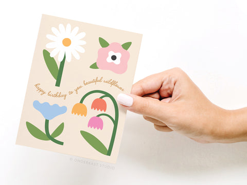 A hand holds the Onderkast Studio “Happy Birthday to You Beautiful Wildflower” greeting card, featuring colorful illustrated flowers and cursive text.