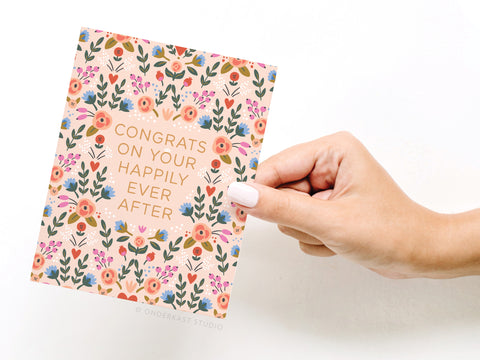 A hand with pale pink nail polish holds the Onderkast Studio Congrats on Your Happily Ever After Floral Greeting Card featuring the greeting in capitals on a white background.
