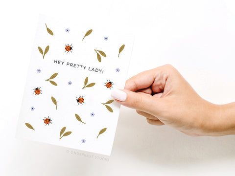 A manicured hand holds the Hey Pretty Lady Ladybugs Greeting Card by Onderkast Studio, featuring vibrant colors, green leaves, blue flowers, and bold “HEY PRETTY LADY!” text for a premium feel.
