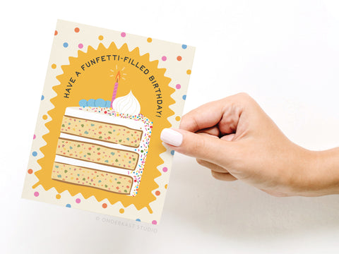 A hand holds the Onderkast Studio Funfetti-filled Birthday Greeting Card, featuring a funfetti cake slice with a candle, sprinkles, and Have a funfetti-filled birthday! text, surrounded by colorful confetti dots.