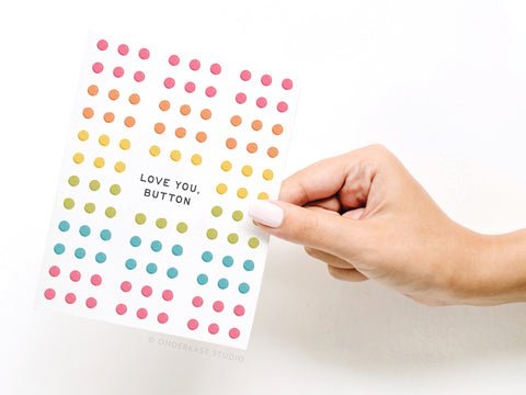 A hand with white-painted nails holds the Love You Button Candy Greeting Card – FINAL STOCK by Onderkast Studio, featuring colorful candy dots and LOVE YOU, BUTTON in bold black text—a nostalgic childhood throwback.