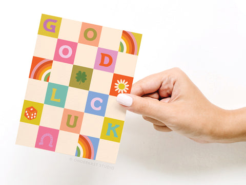 A hand with white nail polish holds the Groovy Good Luck Greeting Card by Onderkast Studio, featuring a checkered “GOOD LUCK” design with charming symbols like a rainbow, clover, daisy, die, and horseshoe.