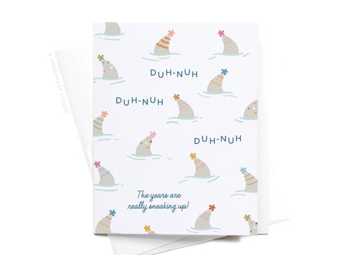 The Shark Fin Party Hats Birthday Greeting Card by Onderkast Studio features cartoon fins in party hats with repeated “DUH-NUH” text and “The years are really sneaking up!”—a playful card for any birthday celebration.