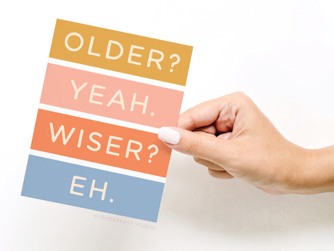 A hand with white painted nails holds the Onderkast Studio Older Not Wiser Birthday Greeting Card, featuring bold white text—OLDER? YEAH. WISER? EH.—on yellow, pink, and blue stripes for a premium birthday greeting.