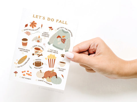 A hand holds the “Let’s Do Fall Greeting Card” by Onderkast Studio, featuring illustrated autumn activities like raking leaves, drinking hot drinks, eating candy, lighting candles, carving pumpkins, and watching football.