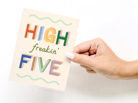 A hand with light pink nail polish holds Onderkast Studio’s High Freakin’ Five Greeting Card, featuring colorful, playful letters on a cream background with wavy green lines—a fun congrats card, made in the USA.