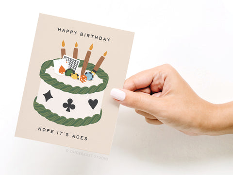 A hand with light nail polish holds the Onderkast Studio Aces Poker Birthday Greeting Card, featuring a cake with candles, playing cards, suits, and the messages “HAPPY BIRTHDAY” and “HOPE IT’S ACES” for a premium birthday touch.