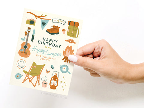 A hand holds the Onderkast Studio Happy Birthday to the Happy Camper Greeting Card, featuring camping-themed illustrations like a guitar, boots, backpack, and lantern. The card says: “Happy Birthday to the Happy Camper. Here’s to another year of adventure.”.