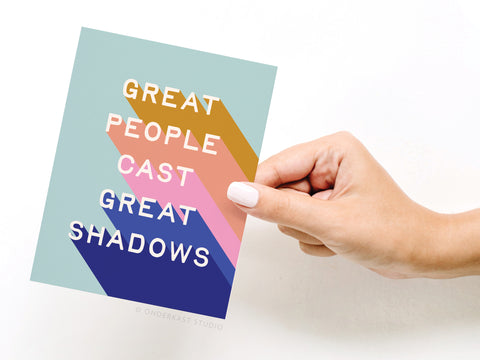 A neatly manicured hand holds the Onderkast Studio Great People Cast Great Shadows Greeting Card, featuring bold text and multicolored diagonal stripes.