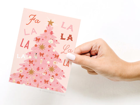 A hand with white nail polish holds the Onderkast Studio Fa La La Pink Christmas Tree Greeting Card featuring a pink tree with ornaments, bows, stars, and Fa La La La La text in various fonts.