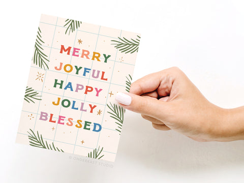 A hand with light pink nail polish holds the Merry Joyful Christmas Spruce Greeting Card by Onderkast Studio, featuring green leaves, gold stars, and bold, colorful MERRY JOYFUL HAPPY JOLLY BLESSED text in vibrant print.