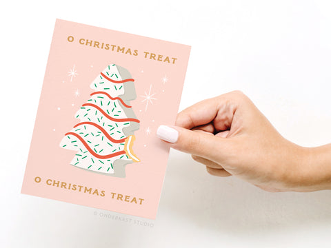 A hand with white nail polish holds the Onderkast Studio O Christmas Treat Greeting Card, showcasing a festive pink card with a Christmas tree cookie, white icing, red and green sprinkles, and the phrase “O CHRISTMAS TREAT.”.