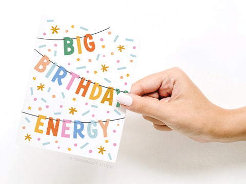 A hand with white-painted nails holds the Onderkast Studio Big Birthday Energy Banner Greeting Card, featuring bold text and festive confetti and star accents on a white background.