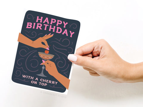 A hand holds the Onderkast Studio Happy Birthday Cocktail With Cherry On Top Greeting Card, featuring two hands placing a cherry on a Manhattan cocktail. The card says Happy Birthday at the top and “With a cherry on top” at the bottom.