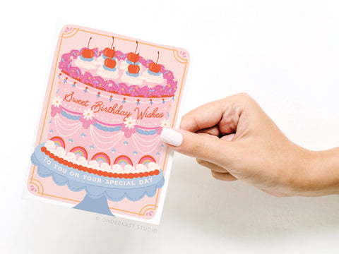 A hand with pale pink nails holds the Onderkast Studio Sweet Birthday Wishes Retro Cake Greeting Card, featuring a colorful tiered cake with cherries and pink accents. The card reads “Sweet Birthday Wishes” and “To You On Your Special Day.”.