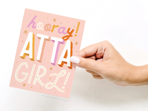 A hand with white nail polish holds the Hooray Atta Girl Greeting Card by Onderkast Studio, featuring colorful, bold hooray! ATTA GIRL! text with stars and playful lettering for a premium look.