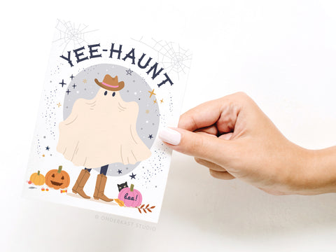 A hand holds the Onderkast Studio Yee-Haunt Greeting Card, featuring a ghost in a cowboy hat and boots with pumpkins, stars, and spider webs—perfect for adding wild west fun to your Halloween theme.
