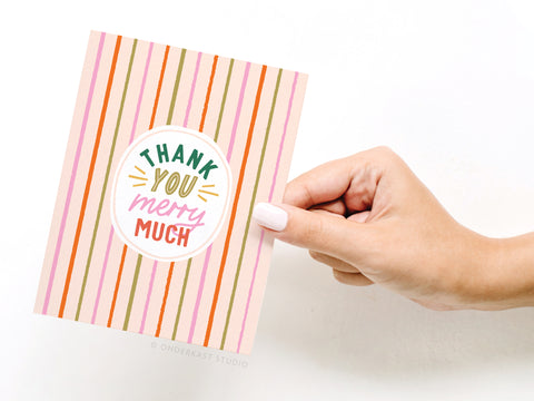 A hand with white nail polish holds the Onderkast Studio Thank You Merry Much Greeting Card, featuring vertical pink, orange, and gold stripes on white and playful lettering—a premium design perfect for holiday cheer.