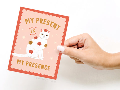 A hand holds the Onderkast Studio My Present Is My Presence Cat Greeting Card, featuring a white cat with red and gold bows and bold, playful text on a vibrant peach and red background.