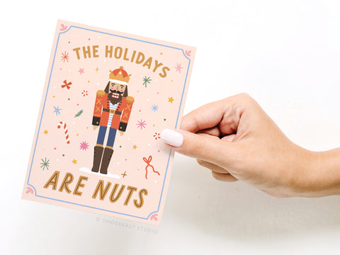 A hand with white nail polish holds Onderkast Studio’s The Holidays Are Nuts Nutcracker Greeting Card, adorned with colorful stars, candy canes, and confetti. The card adds humor to the season, boldly stating: The holidays are nuts.