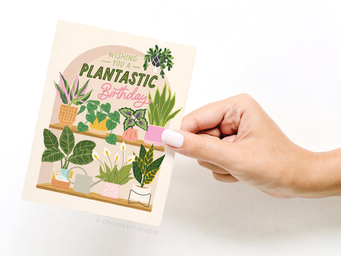 A hand with white nail polish holds the Onderkast Studio Wishing You a Plantastic Birthday Greeting Card, featuring illustrated houseplants on shelves and the cheerful birthday message.