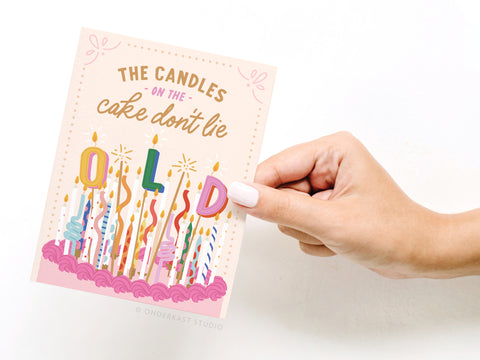 A hand with white nail polish holds the Onderkast Studio The Candles on the Cake Don’t Lie Greeting Card, featuring a cake with many lit candles spelling “O L D” and the caption, “The candles on the cake don’t lie.”.