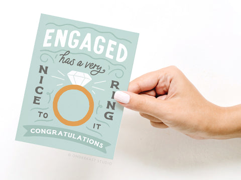 A hand with white-painted nails holds the Onderkast Studio Engaged Has a Very Nice Ring to It greeting card, featuring a diamond ring illustration and congratulatory message.