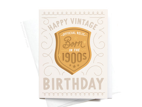 The Onderkast Studio Born in the 1900s Vintage Birthday Greeting Card features a gold badge reading Official Relic—Born in the 1900s and Happy Vintage Birthday in beige and gold, accented with decorative swirls.