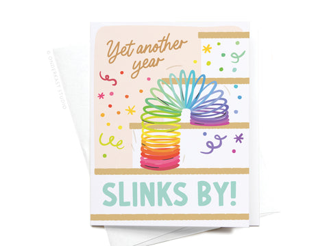 The Yet Another Year Slinks By Greeting Card by Onderkast Studio features a rainbow Slinky on stairs with confetti, playful Yet another year SLINKS BY! text, and comes with an envelope—perfect for nostalgic birthday wishes.