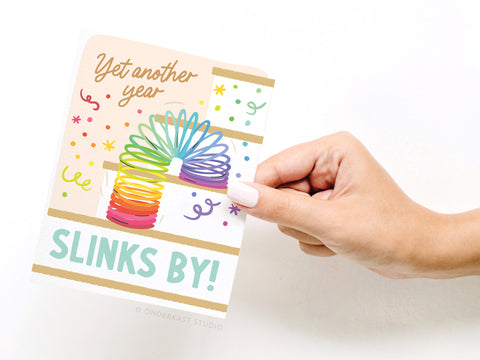 A hand holds the Onderkast Studio Yet Another Year Slinks By Greeting Card, featuring a rainbow Slinky toy, confetti, and playful fonts—perfect for nostalgic birthday wishes.