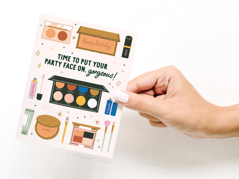 A hand holds the Time to Put Your Party Face On Greeting Card by Onderkast Studio, featuring makeup illustrations, a birthday cake, and festive text—perfect for makeup lovers birthdays. A Happy Birthday insert appears in the background.