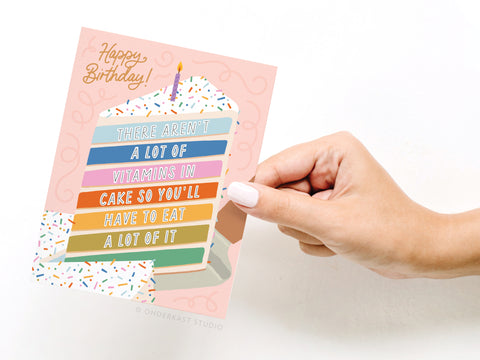 A hand holds Onderkast Studio’s Not a Lot of Vitamins in Cake Greeting Card, featuring a colorful cake slice and the message: “Happy Birthday! There aren’t a lot of vitamins in cake, so you’ll have to eat a lot of it.”.
