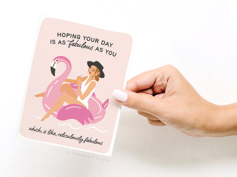 A hand holds the Onderkast Studio Fabulous Flamingo Floatie Greeting Card, showing a woman in a hat and swimsuit on a pink flamingo float, sipping a drink. The card says, Hoping your day is as fabulous as you! Printed in the USA.