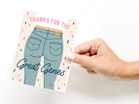 A light-skinned hand with white nails holds the “Thanks for the Great Genes Mom Jeans Greeting Card” by Onderkast Studio, featuring illustrated jeans and colorful sprinkles. Printed in the USA.