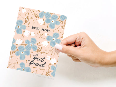 A hand with light pink nail polish holds the Onderkast Studio Best Mom Best Friend Greeting Card, featuring blue and white flowers on a beige background—perfect for Mothers Day gifting.