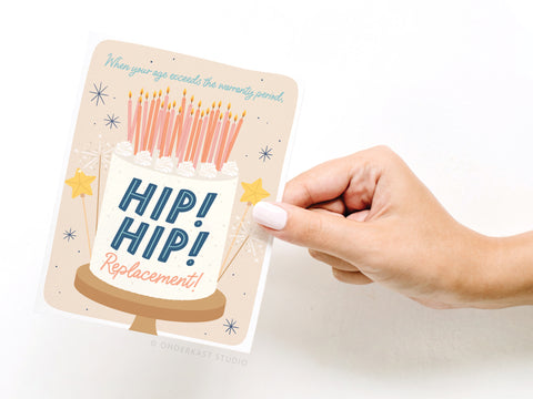 A hand holds the Hip Hip Replacement Greeting Card by Onderkast Studio, featuring a cake with many candles and the message: When your age exceeds the warranty period, HIP! HIP! Replacement! Ideal birthday humor for any celebration.