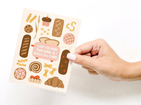 A hand holds the Onderkast Studio I Love You More Than Carbs Greeting Card, featuring charming bread and pastry illustrations and the message: “I love you more than carbs, & I really love carbs.”.