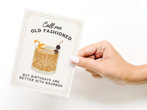 A hand with white nail polish holds the Onderkast Studio Call Me Old Fashioned Cocktail Greeting Card featuring a cocktail illustration and the message: Call me old fashioned but birthdays are better with bourbon—a perfect mixology birthday card.