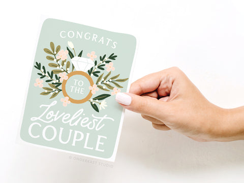 A hand with white nail polish holds the Onderkast Studio Congrats to the Loveliest Couple Greeting Card, featuring floral designs and a diamond ring on a pastel green background. Printed in the USA.