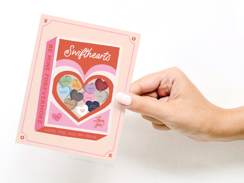A hand holds the Swifthearts Candy Box Greeting Card by Onderkast Studio, featuring heart-shaped candies with phrases from Taylors eras like Lover, Bejeweled, and Red. The border reads “Be Mine Forevermore”—a sweet gift for any Swiftie.