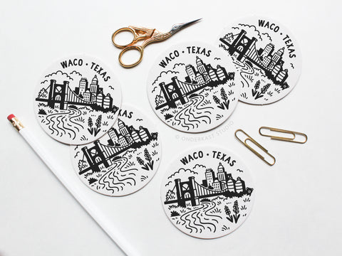 Five round Waco Texas Skyline Letterpress Coasters by Onderkast Studio are arranged on a white surface with gold scissors, a white pencil with a pink eraser, and two gold paper clips nearby.