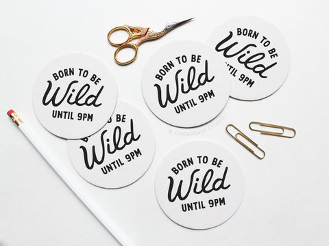 A white pencil, gold scissors, and two paperclips sit beside the Onderkast Studio Born To Be Wild Until 9pm Letterpress Coaster Set—five round white coasters with bold black text, packaged in a recyclable kraft box.