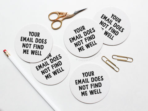 The Your Email Does Not Find Me Well Letterpress Coaster Set by Onderkast Studio features black text on round coasters, styled with a white pencil, gold scissors, and two gold paperclips on a white background.