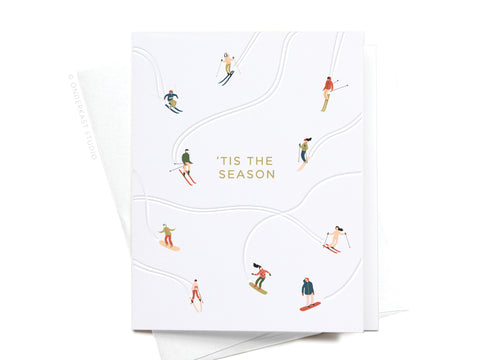 The ‘Tis the Season Skiiers Letterpress Greeting Card by Onderkast Studio features small illustrated skiers on white slopes with curved trails and gold “‘TIS THE SEASON” text in the center. An envelope is partially visible behind the card.