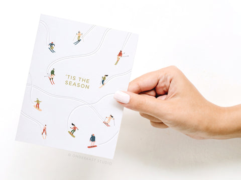 A hand with light nail polish holds the Tis the Season Skiiers Letterpress Greeting Card by Onderkast Studio, featuring small skiers on white slopes and gold TIS THE SEASON text—perfect for festive greetings.