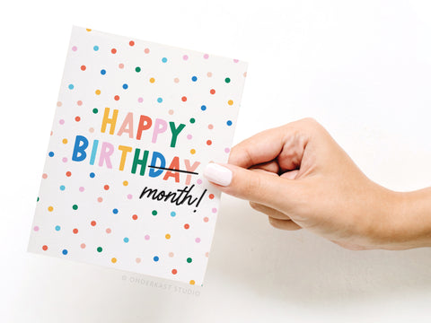 A hand with light pink nail polish holds the Onderkast Studio Happy Birth-month Letterpress Greeting Card, featuring colorful polka dots and bold, multicolored HAPPY BIRTHDAY month! text for a joyful birth-month celebration.