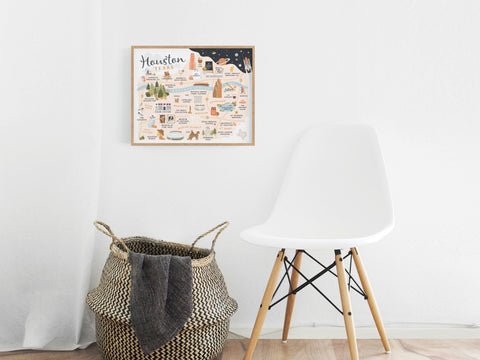 A modern white chair and a woven basket with a dark blanket sit against a white wall, topped by the Houston Map Art Print from Onderkast Studio, which showcases illustrated neighborhoods and local landmarks.